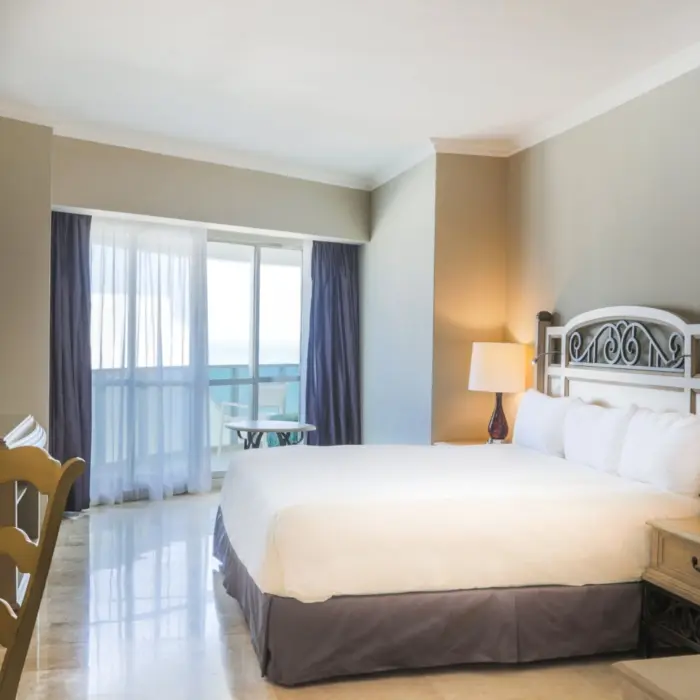 A spacious hotel suite with a king-sized bed, marble floors, a desk, and balcony access to a sea view.
