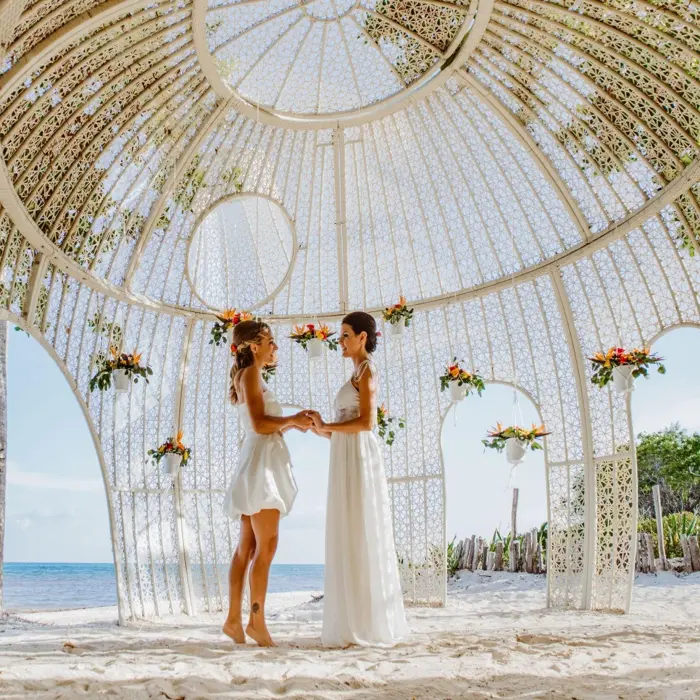 Two brides holding hands in a beautiful white gazebo wedding venue on a sunny beach.