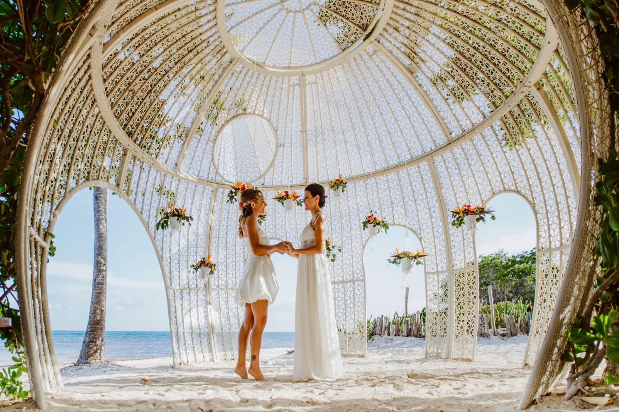 Two brides holding hands in a beautiful white gazebo wedding venue on a sunny beach.