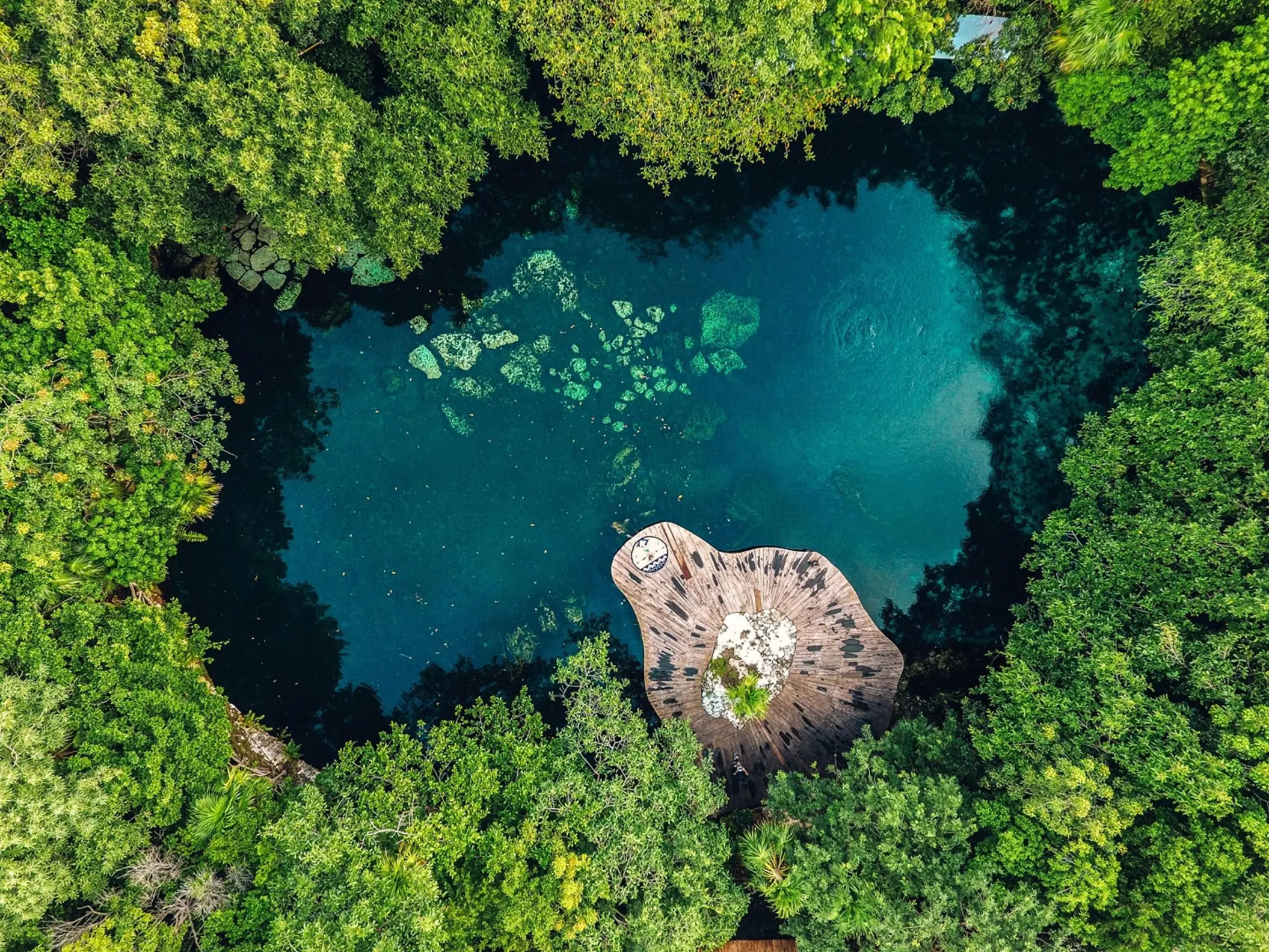 An aerial view of a vibrant blue cenote surrounded by dense green trees at Sandos Caracol Eco Resort.