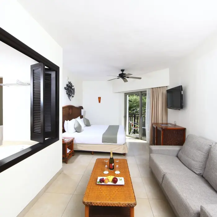 A bright deluxe select suite with a bed, sofa, coffee table with fruit and champagne, and a balcony.