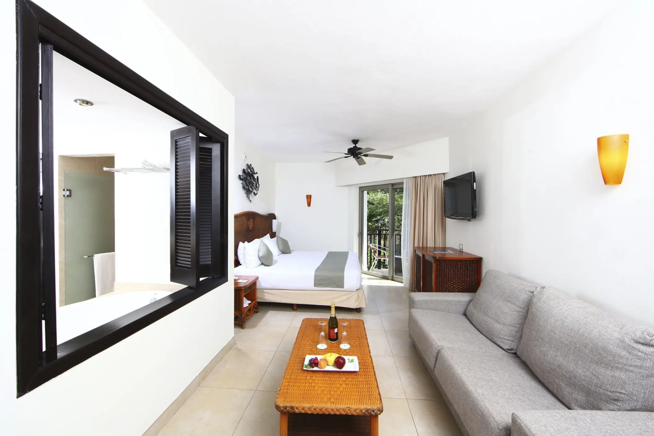 A bright deluxe select suite with a bed, sofa, coffee table with fruit and champagne, and a balcony.