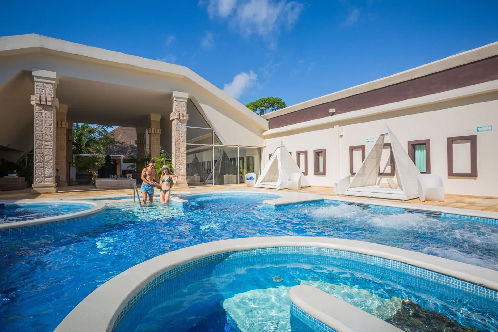 A couple enjoys a large, inviting pool area with cabanas and modern architecture at Sandos Caracol.