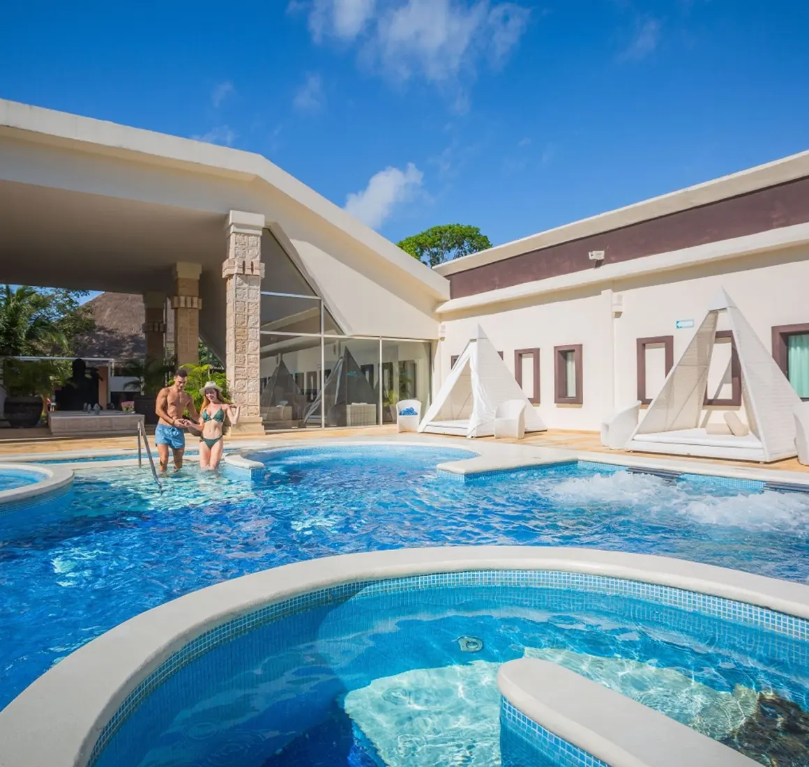 A couple enjoys a large, inviting pool area with cabanas and modern architecture at Sandos Caracol.
