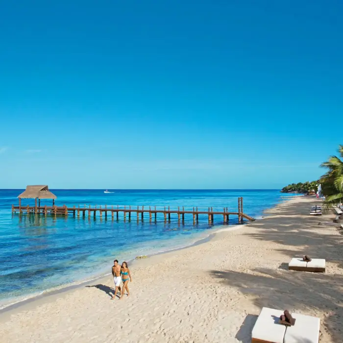 White-sand beach lined with thatched umbrellas and palm trees at an adults-only Riviera Maya resort.