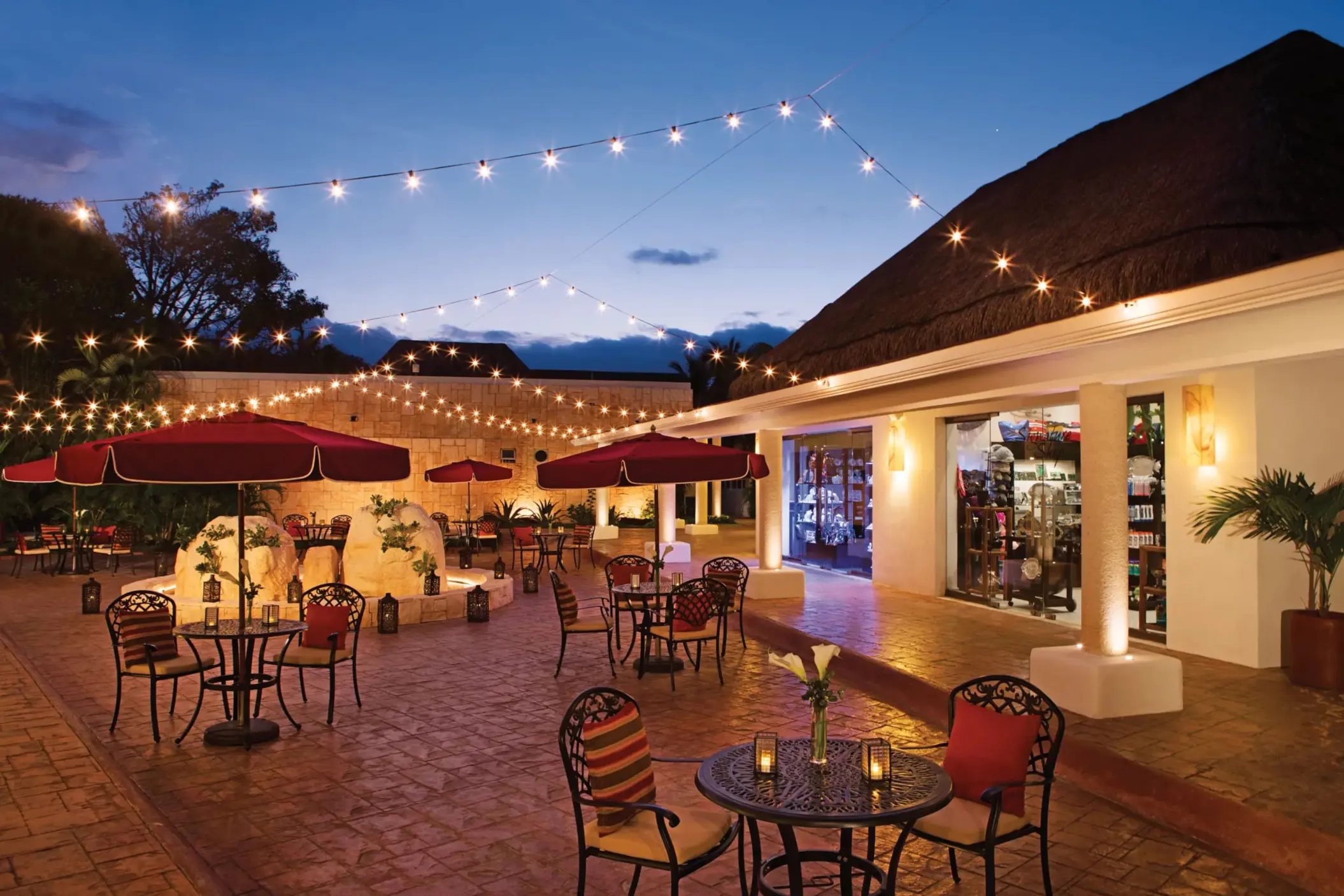 Evening lights strung above a charming open-air plaza with patio seating and boutique-style shops.