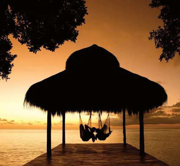 Couple relaxing in hammocks under a palapa at sunset with golden sky over the calm ocean.