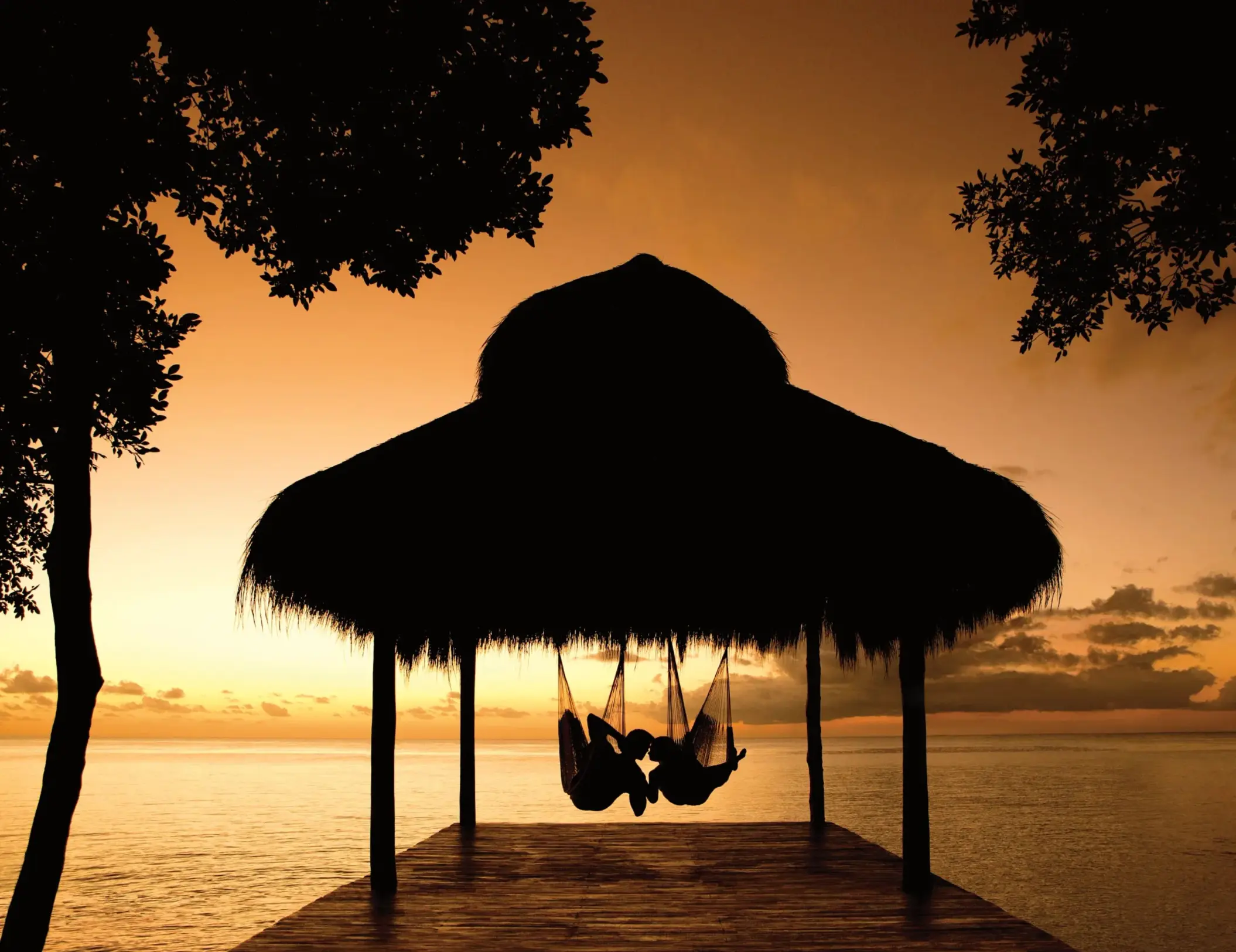 Couple relaxing in hammocks under a palapa at sunset with golden sky over the calm ocean.