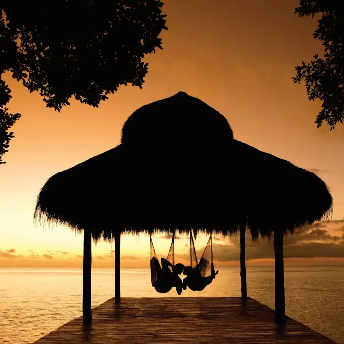 Couple relaxing in hammocks under a palapa at sunset with golden sky over the calm ocean.