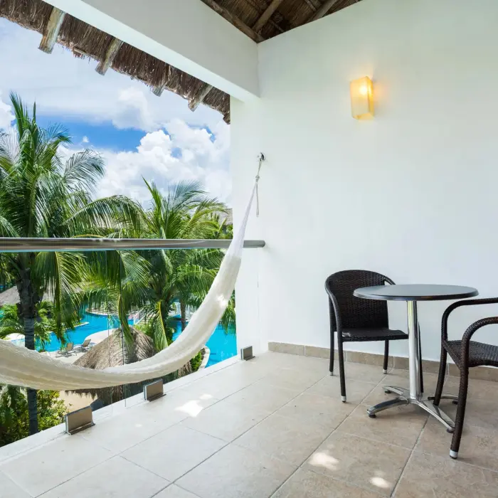 Balcony with pool view, hammock, and two wicker chairs beside a small table under a thatched roof.