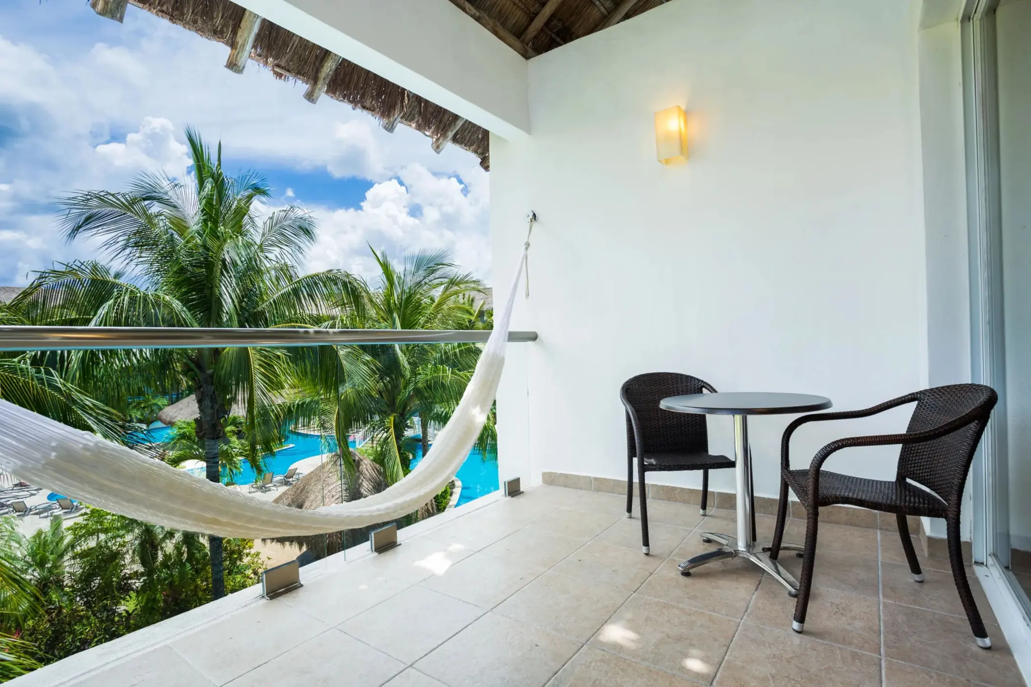 Balcony with pool view, hammock, and two wicker chairs beside a small table under a thatched roof.