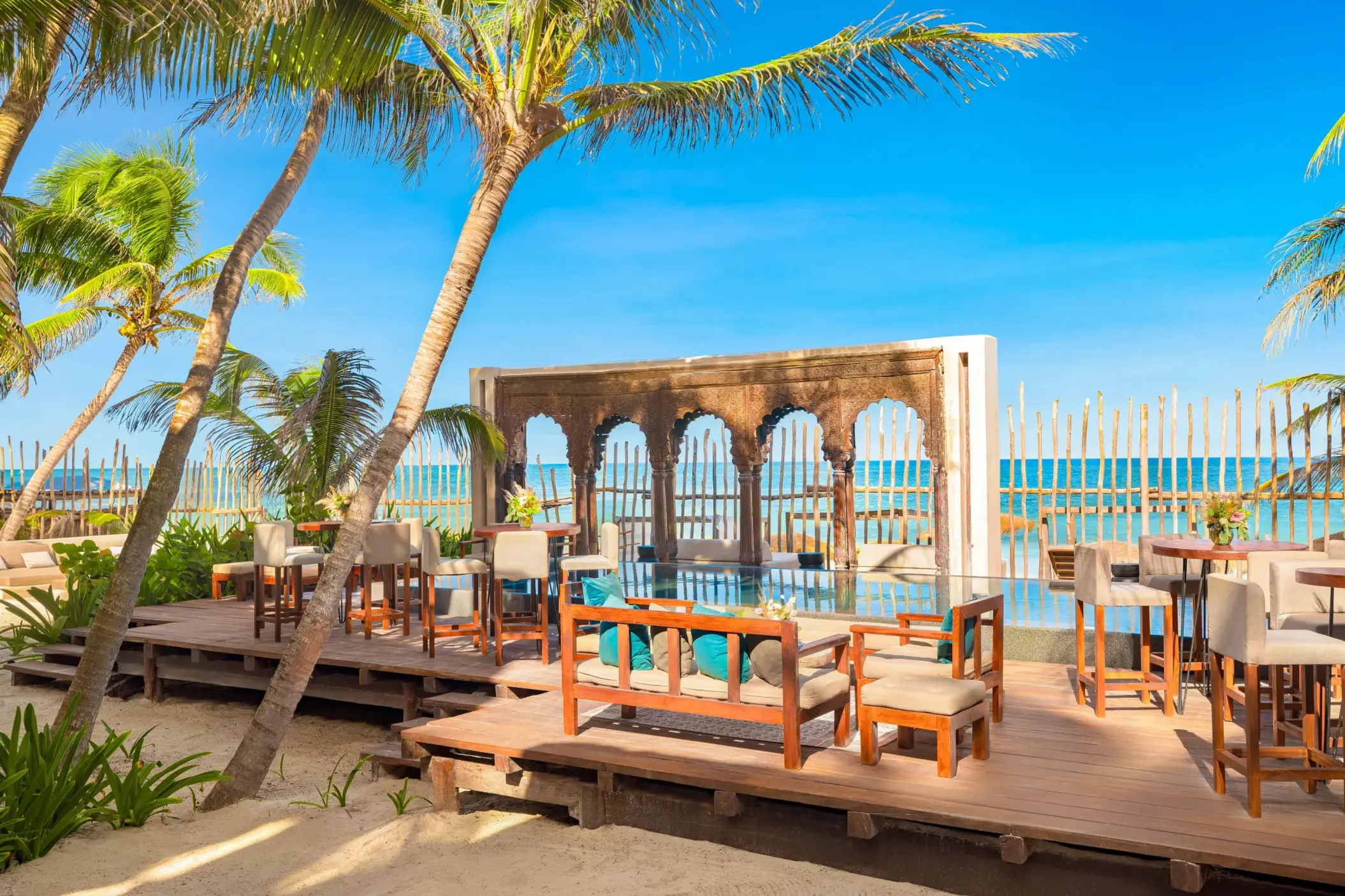 Outdoor beachfront lounge with wooden deck, tropical palms, and rustic arches framing the ocean view.