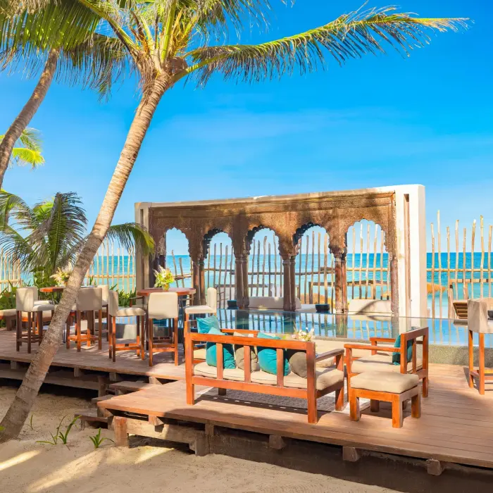 Outdoor beachfront lounge with wooden deck, tropical palms, and rustic arches framing the ocean view.