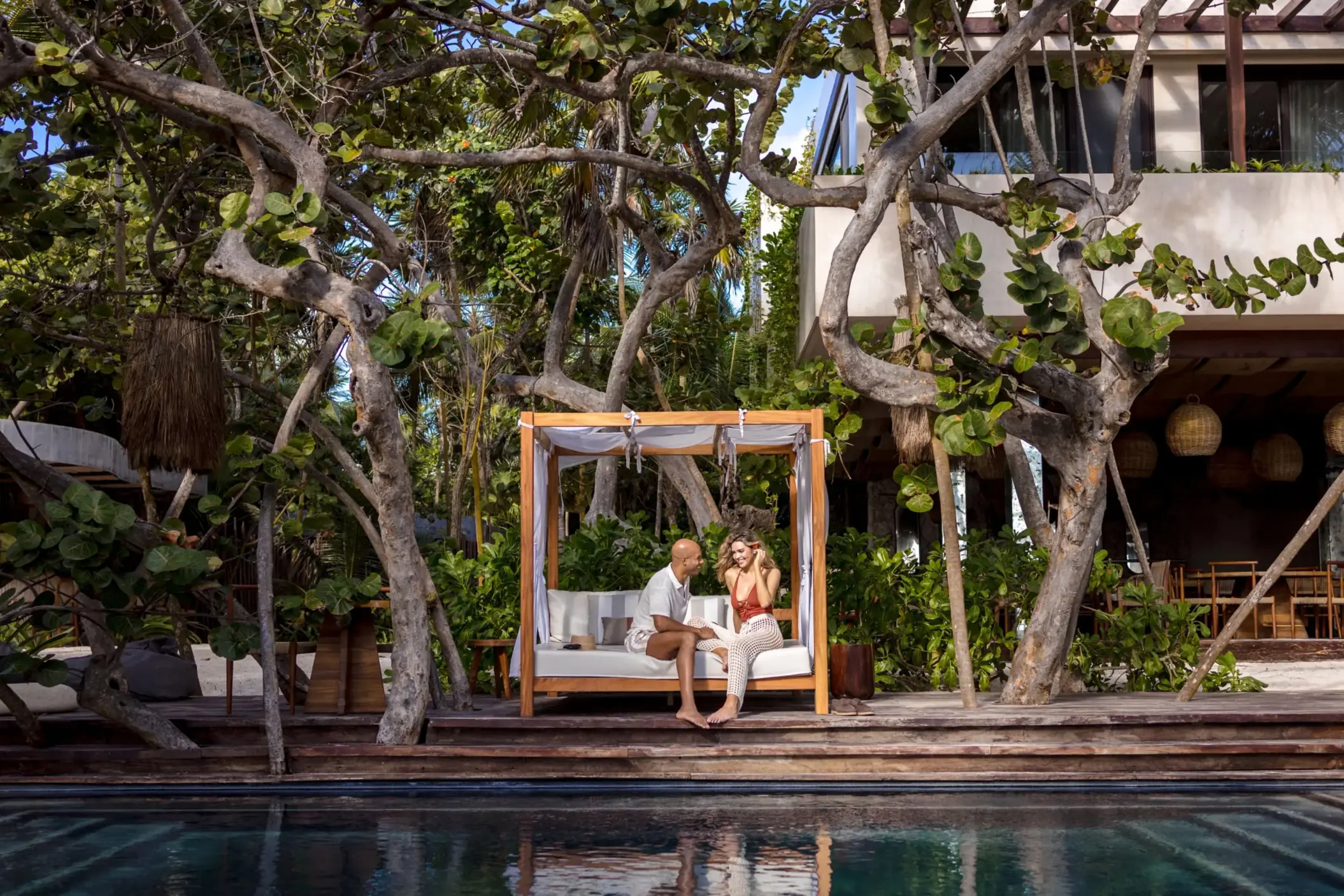 Couple relaxing on a canopy bed by the pool, surrounded by tropical trees and rustic wooden decking.