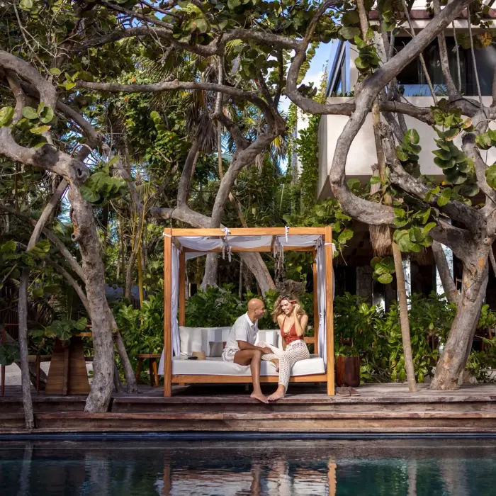 Couple relaxing on a canopy bed by the pool, surrounded by tropical trees and rustic wooden decking.