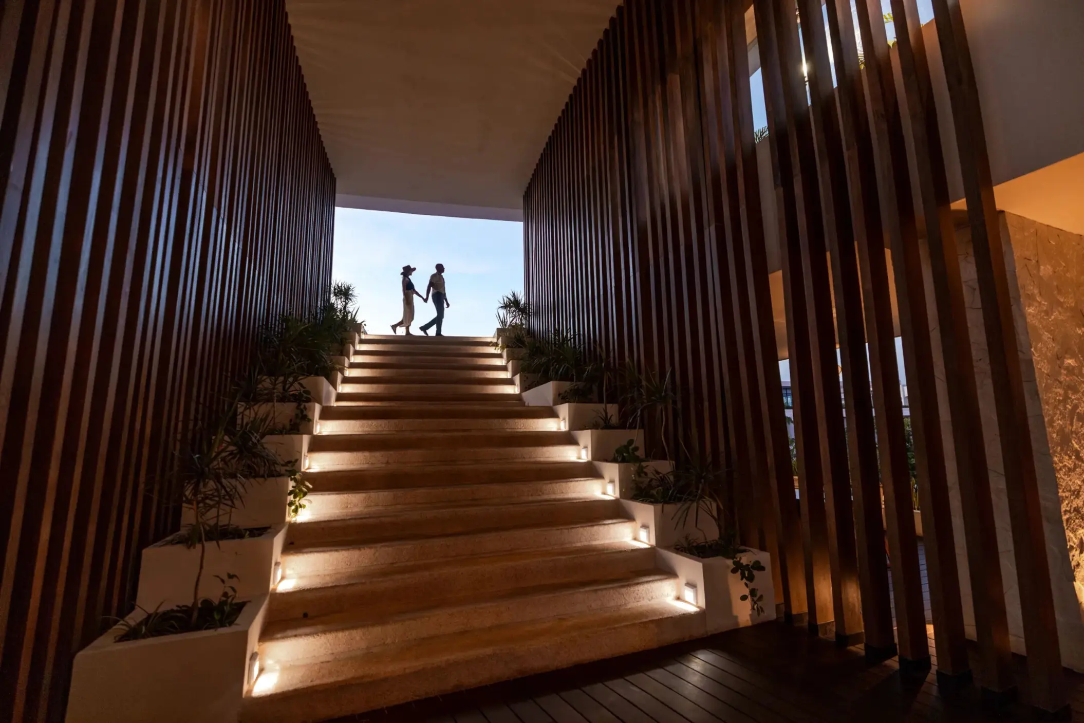 Silhouetted couple walking up illuminated stairs framed by modern wood paneling and soft lighting.
