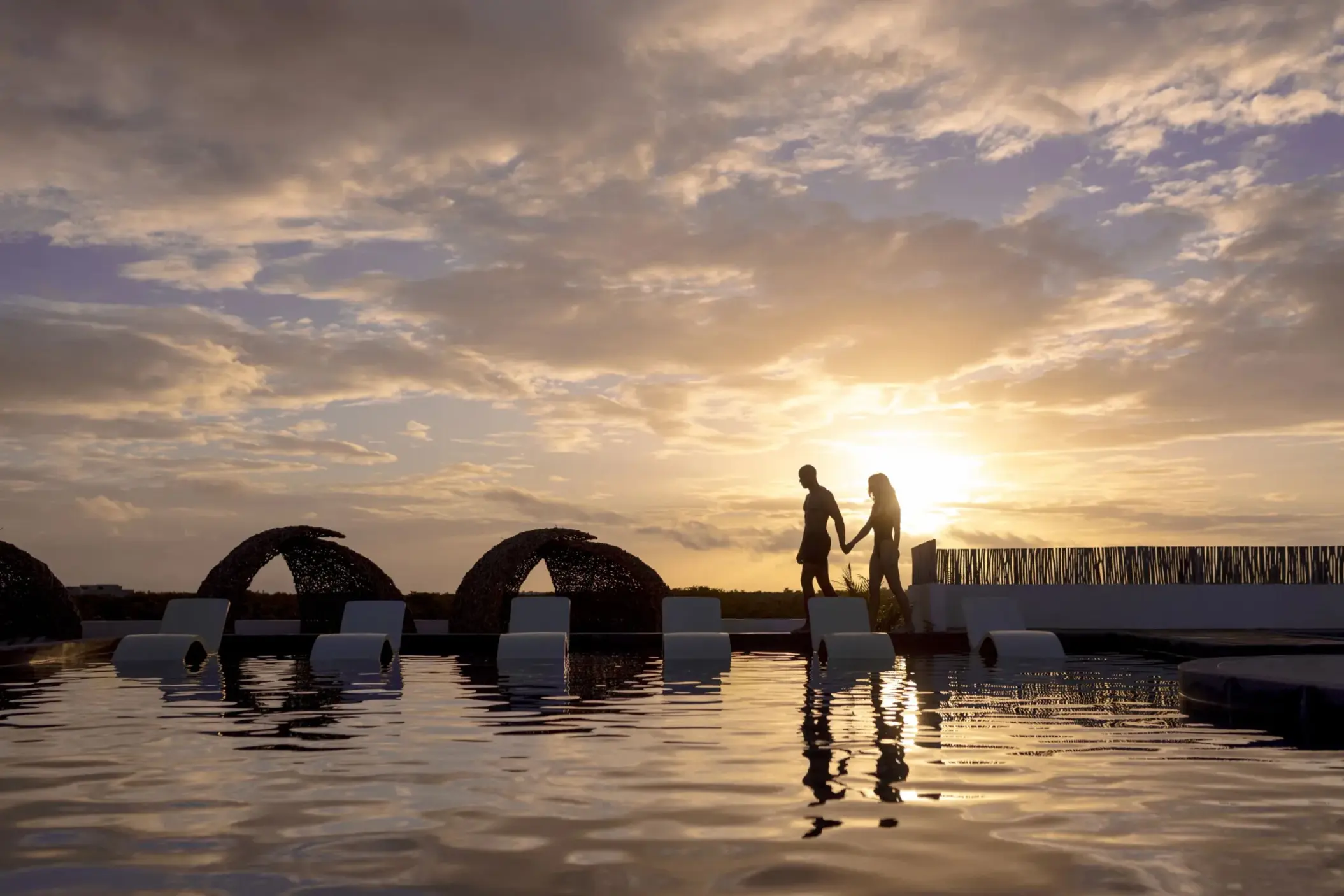Couple walking hand in hand by the pool at sunset with lounge chairs and canopy beds in the background.