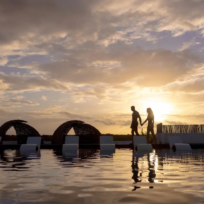Couple walking hand in hand by the pool at sunset with lounge chairs and canopy beds in the background.