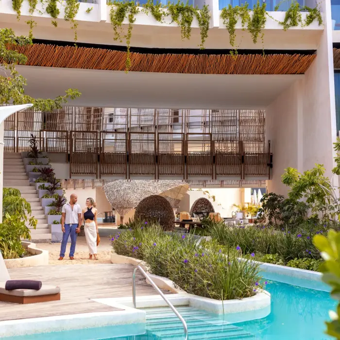 Couple walking near a serene pool with modern architecture, greenery, and outdoor loungers nearby.