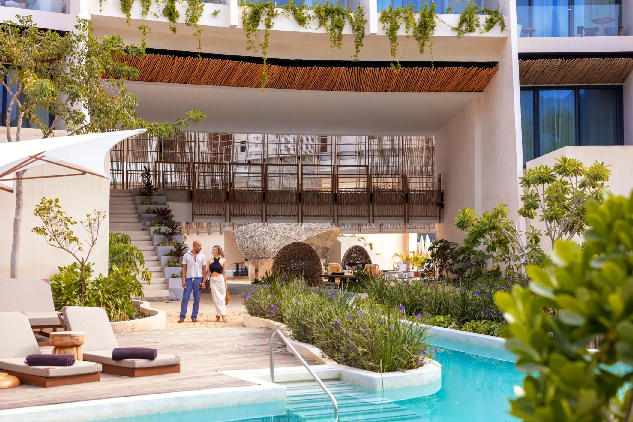 Couple walking near a serene pool with modern architecture, greenery, and outdoor loungers nearby.