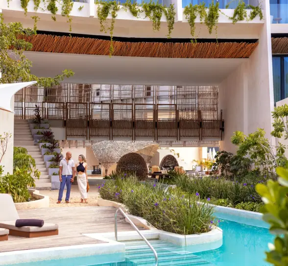 Couple walking near a serene pool with modern architecture, greenery, and outdoor loungers nearby.