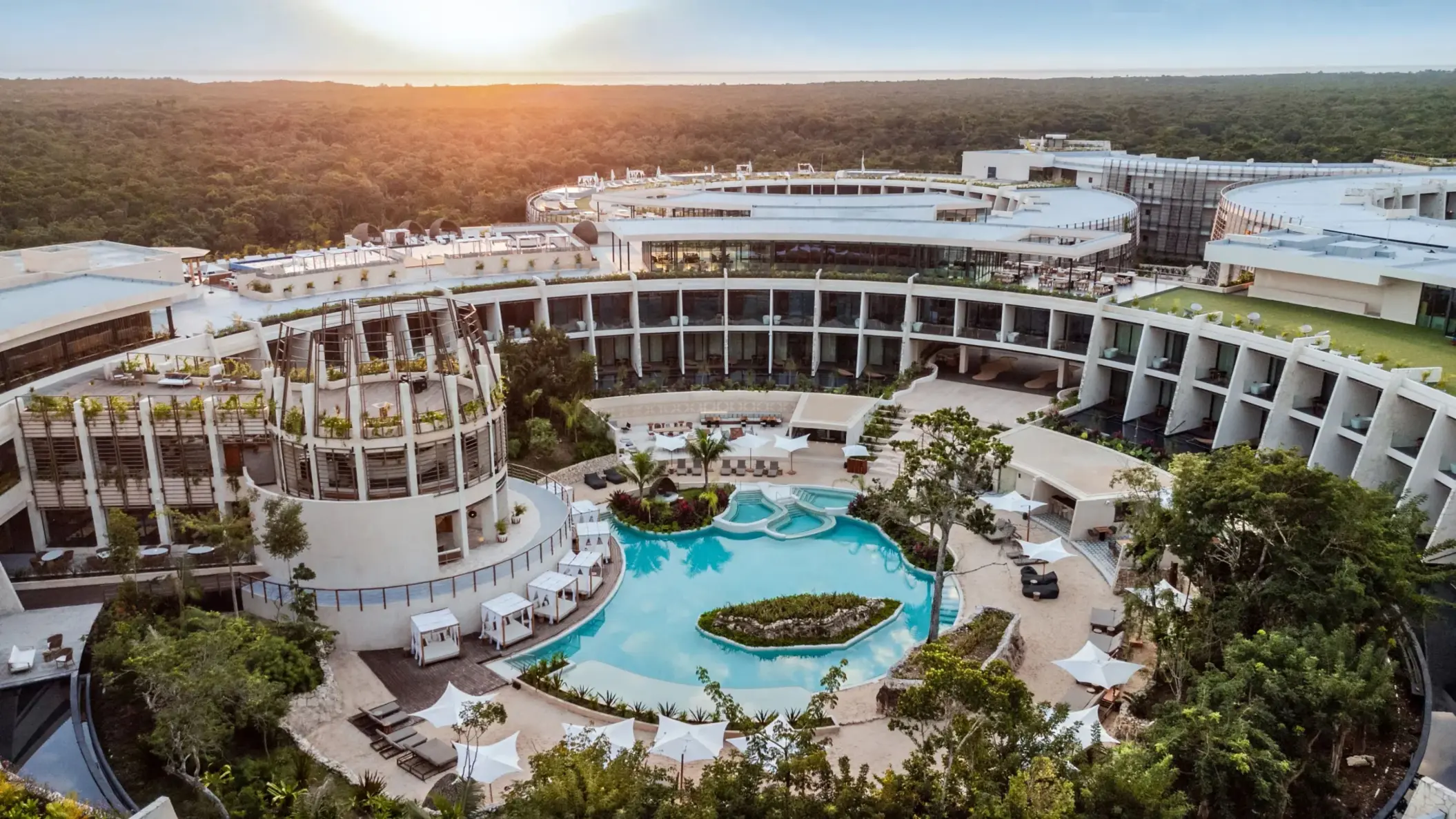 Aerial view of resort architecture with circular buildings, lush greenery, and a lagoon-style pool at sunset.