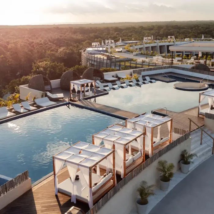 Rooftop infinity pool with white cabanas, loungers, and jungle views under golden sunset light.