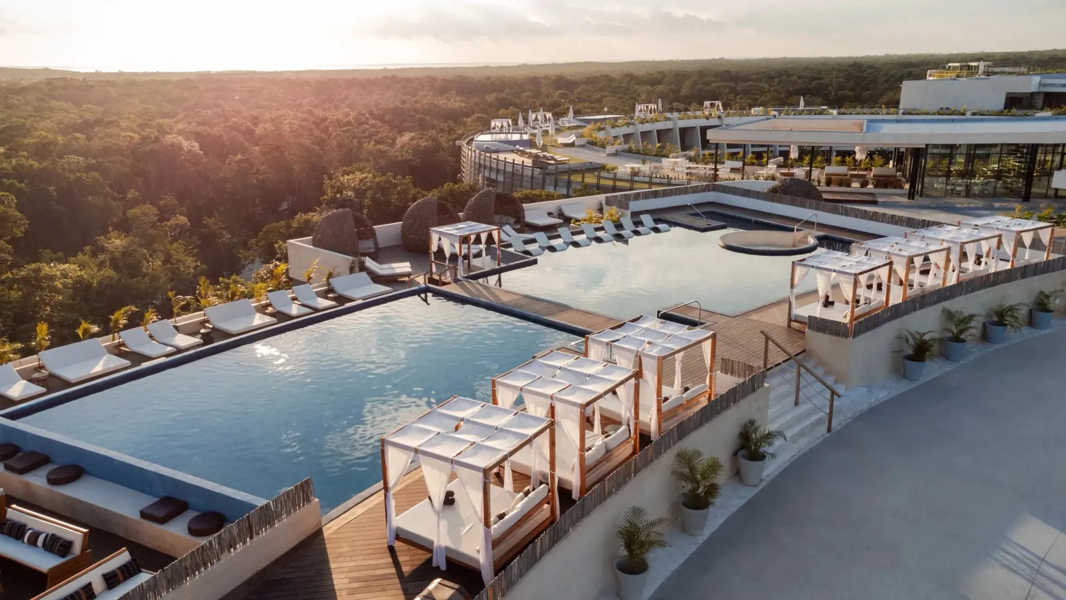 Rooftop infinity pool with white cabanas, loungers, and jungle views under golden sunset light.