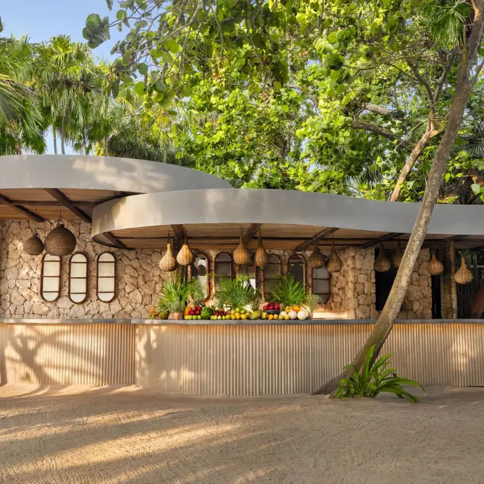 Tropical beach bar with stone walls, hanging lamps, and a vibrant fruit display under lush jungle canopy.