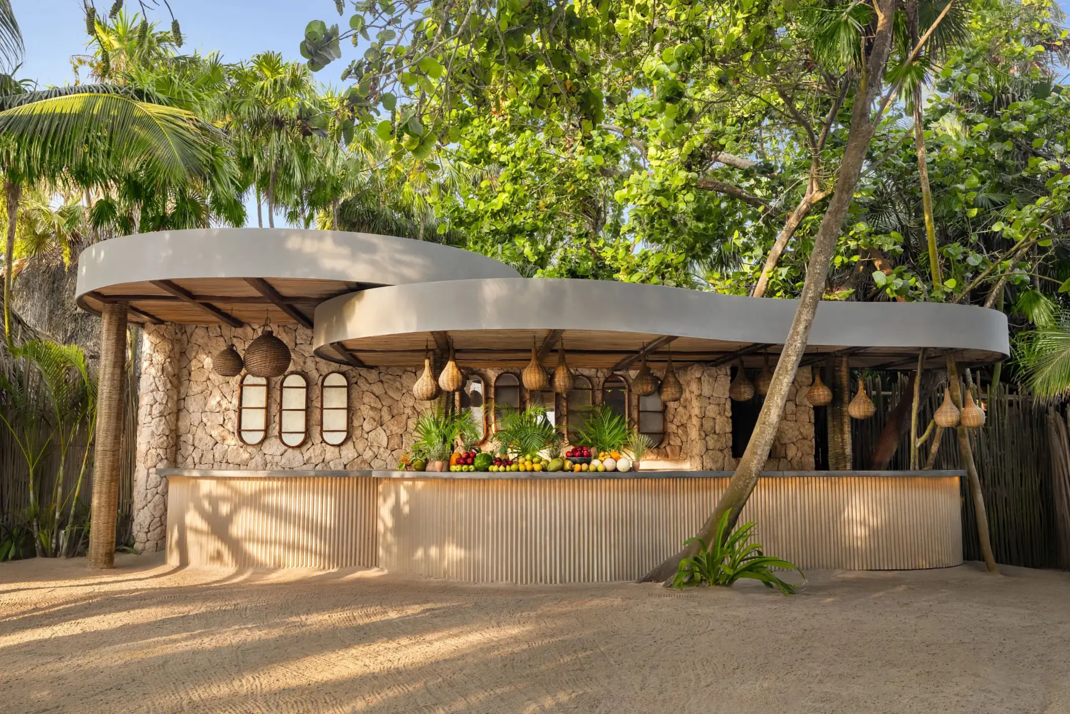 Tropical beach bar with stone walls, hanging lamps, and a vibrant fruit display under lush jungle canopy.