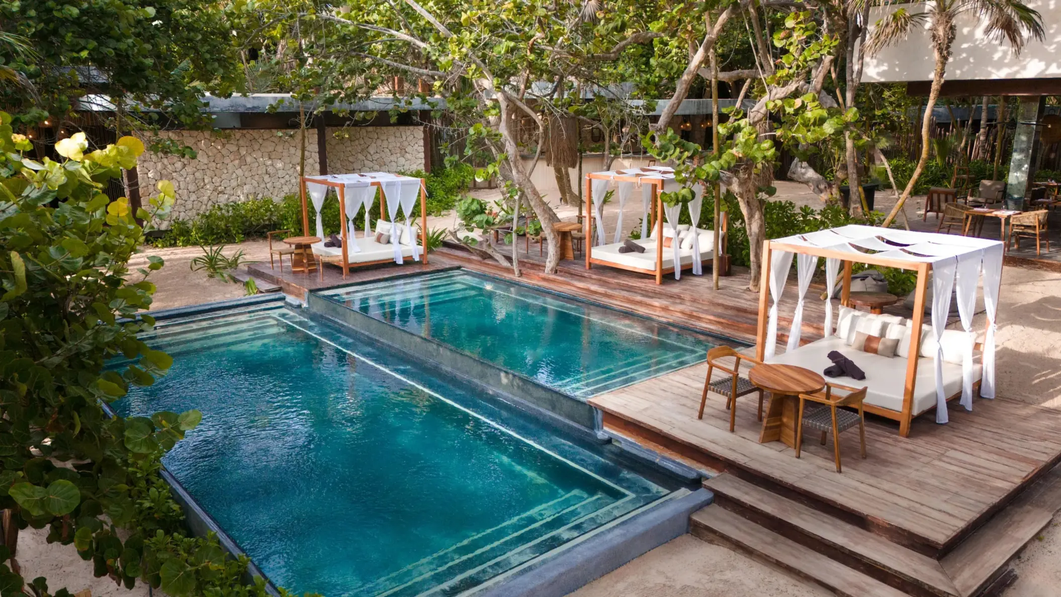 Lush beach club pool with sun loungers, shaded pergolas, and palm trees surrounding a serene swimming area.