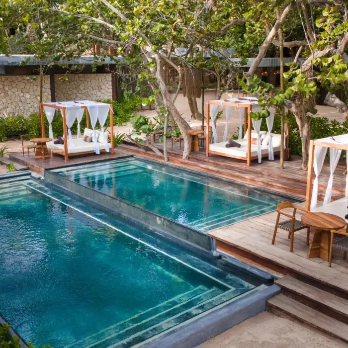 Lush beach club pool with sun loungers, shaded pergolas, and palm trees surrounding a serene swimming area.