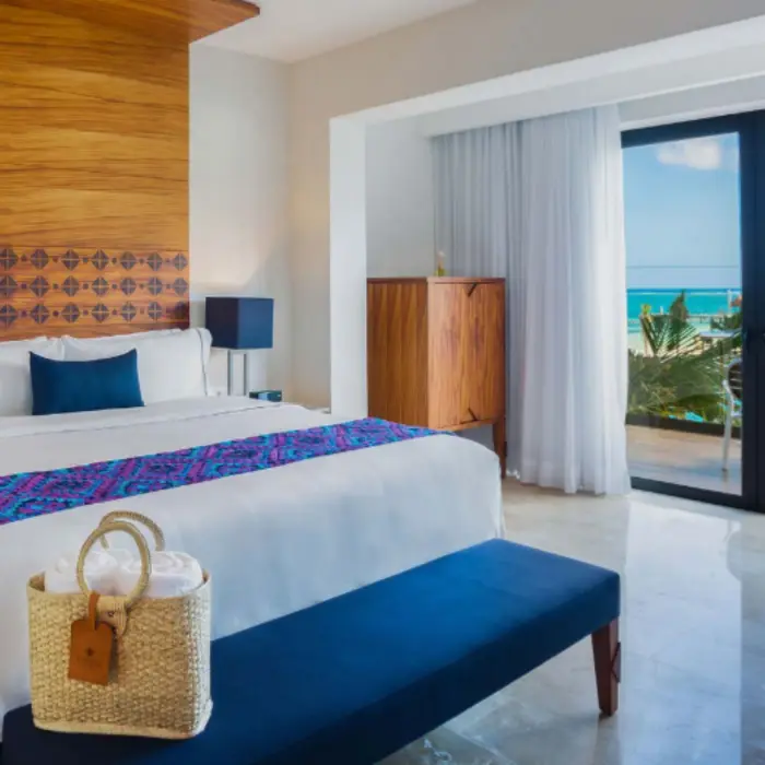 Elegant hotel room with king bed, ocean view balcony, wood accents, and natural light flooding through glass doors.