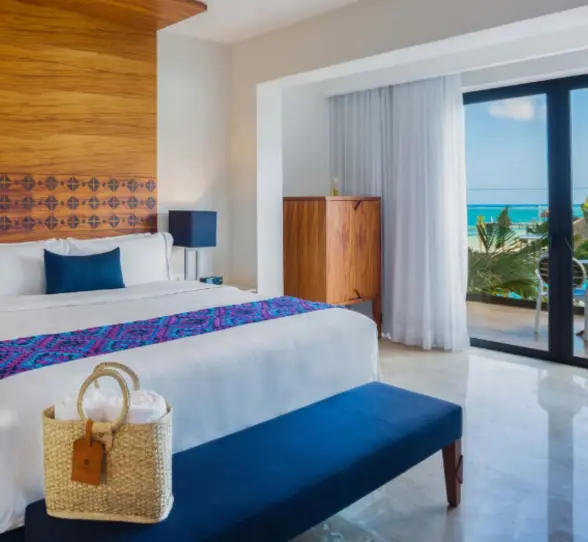 Elegant hotel room with king bed, ocean view balcony, wood accents, and natural light flooding through glass doors.