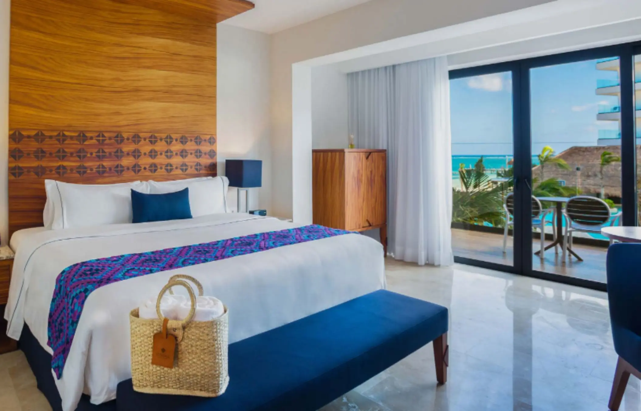 Elegant hotel room with king bed, ocean view balcony, wood accents, and natural light flooding through glass doors.