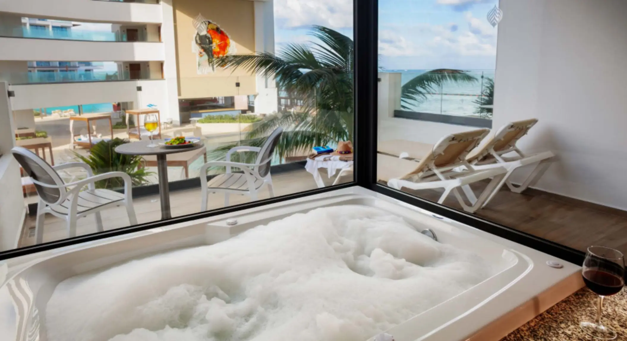 Foamy jacuzzi tub beside a floor-to-ceiling window with ocean views, patio seating, and sun loungers outside.