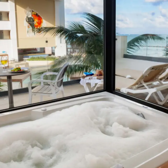 Foamy jacuzzi tub beside a floor-to-ceiling window with ocean views, patio seating, and sun loungers outside.
