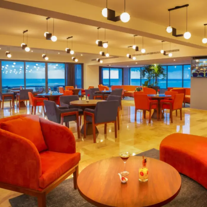Chic oceanview lounge with bold orange seating, modern lighting, and cozy tables beside large glass windows.