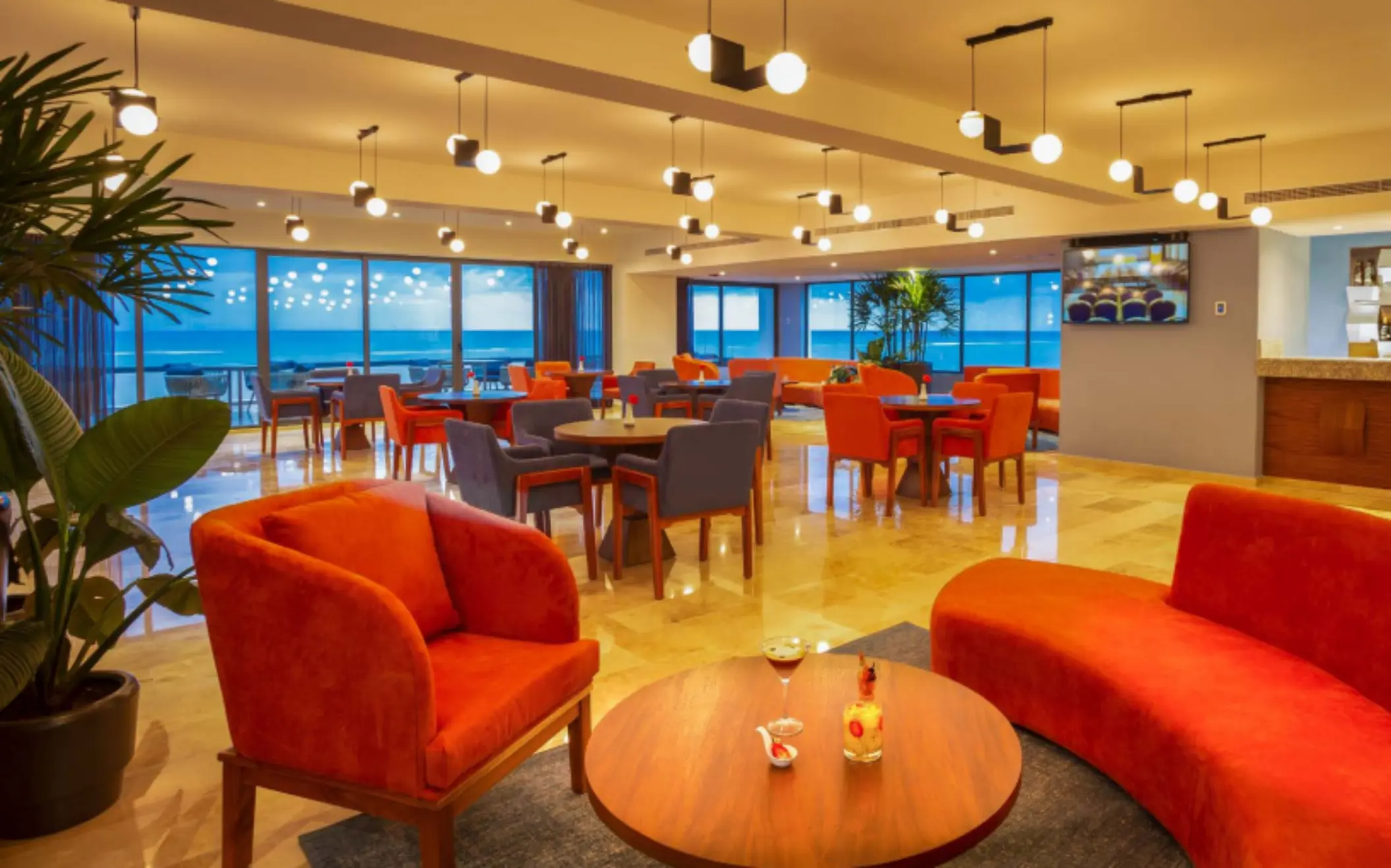 Chic oceanview lounge with bold orange seating, modern lighting, and cozy tables beside large glass windows.