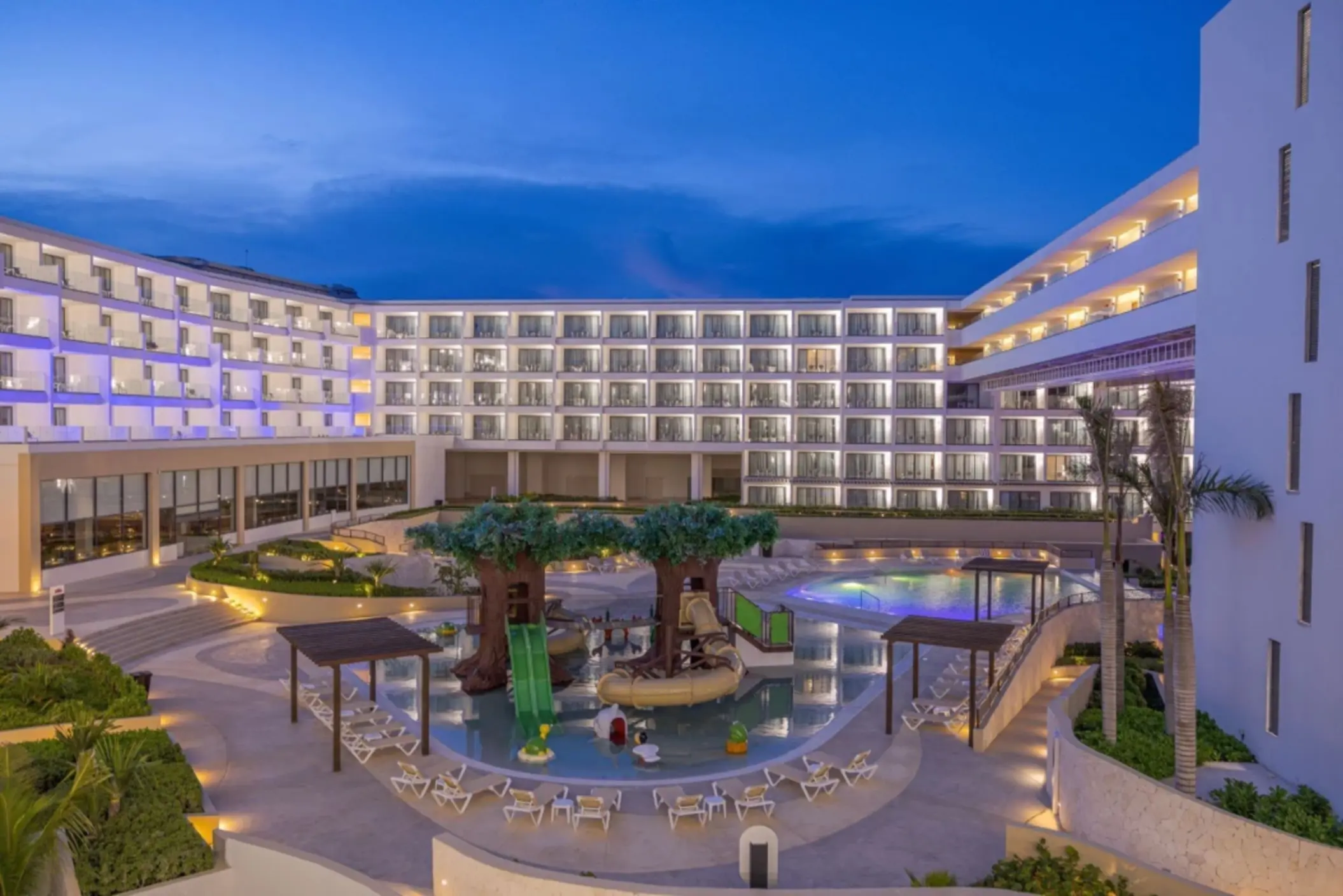Evening view of kids’ splash zone with tree slides and illuminated hotel balconies surrounding the pool.