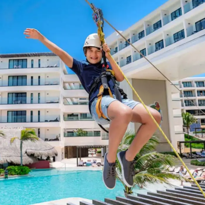 A child ziplining over the pool area at a modern beachfront resort with clear skies and palm trees in the background.