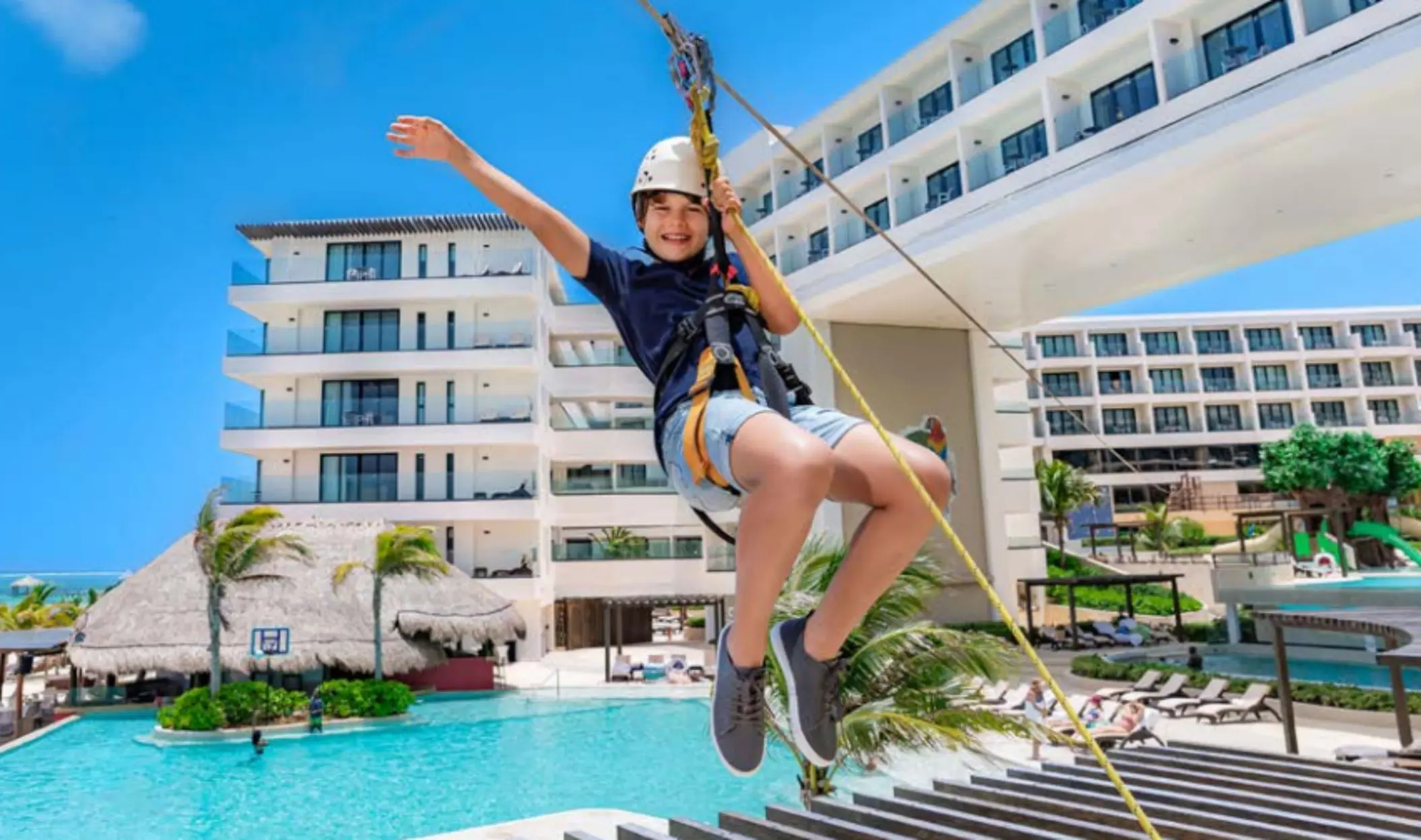 A child ziplining over the pool area at a modern beachfront resort with clear skies and palm trees in the background.