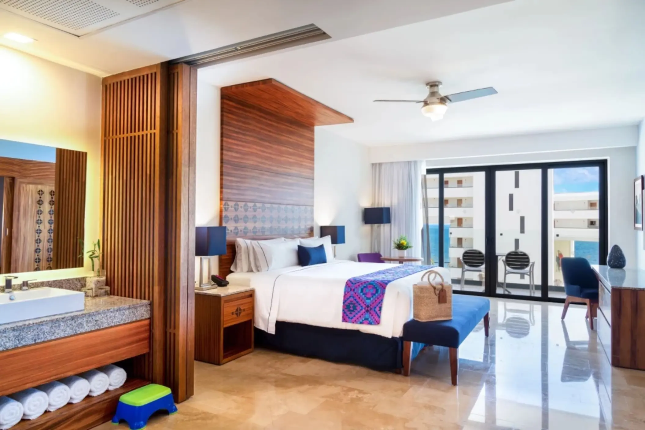 Modern oceanview room with king bed, colorful runner, balcony seating, and open vanity with granite countertop.