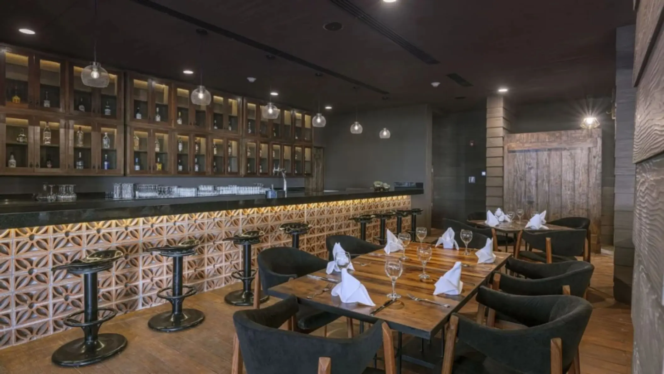 Rustic-style indoor bar with wood cabinets, hanging lights, tiled bar front, and intimate dining setup with dark chairs.
