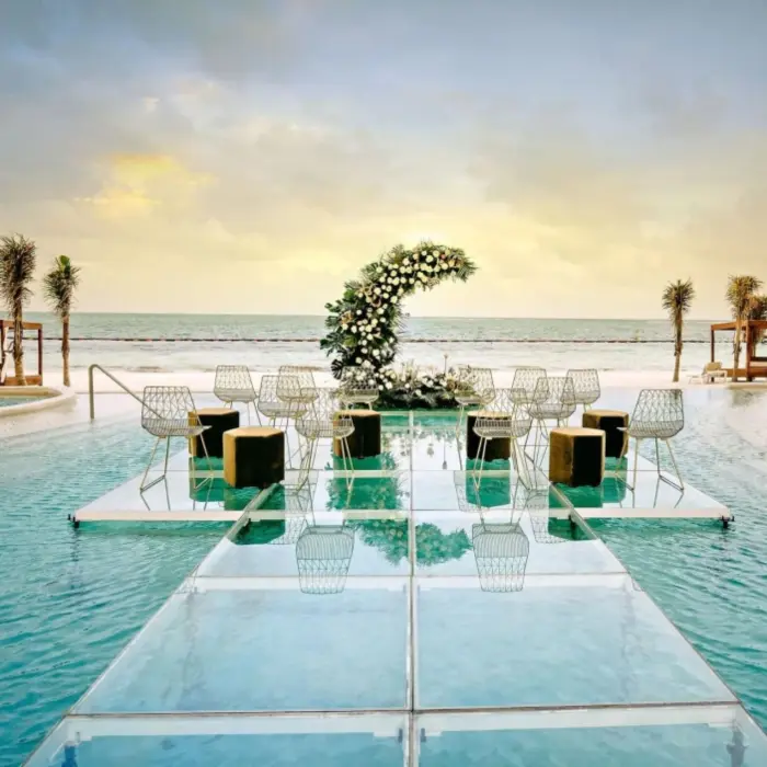 Floating wedding ceremony platform over a pool with ocean backdrop, floral arch, and modern wire chairs.