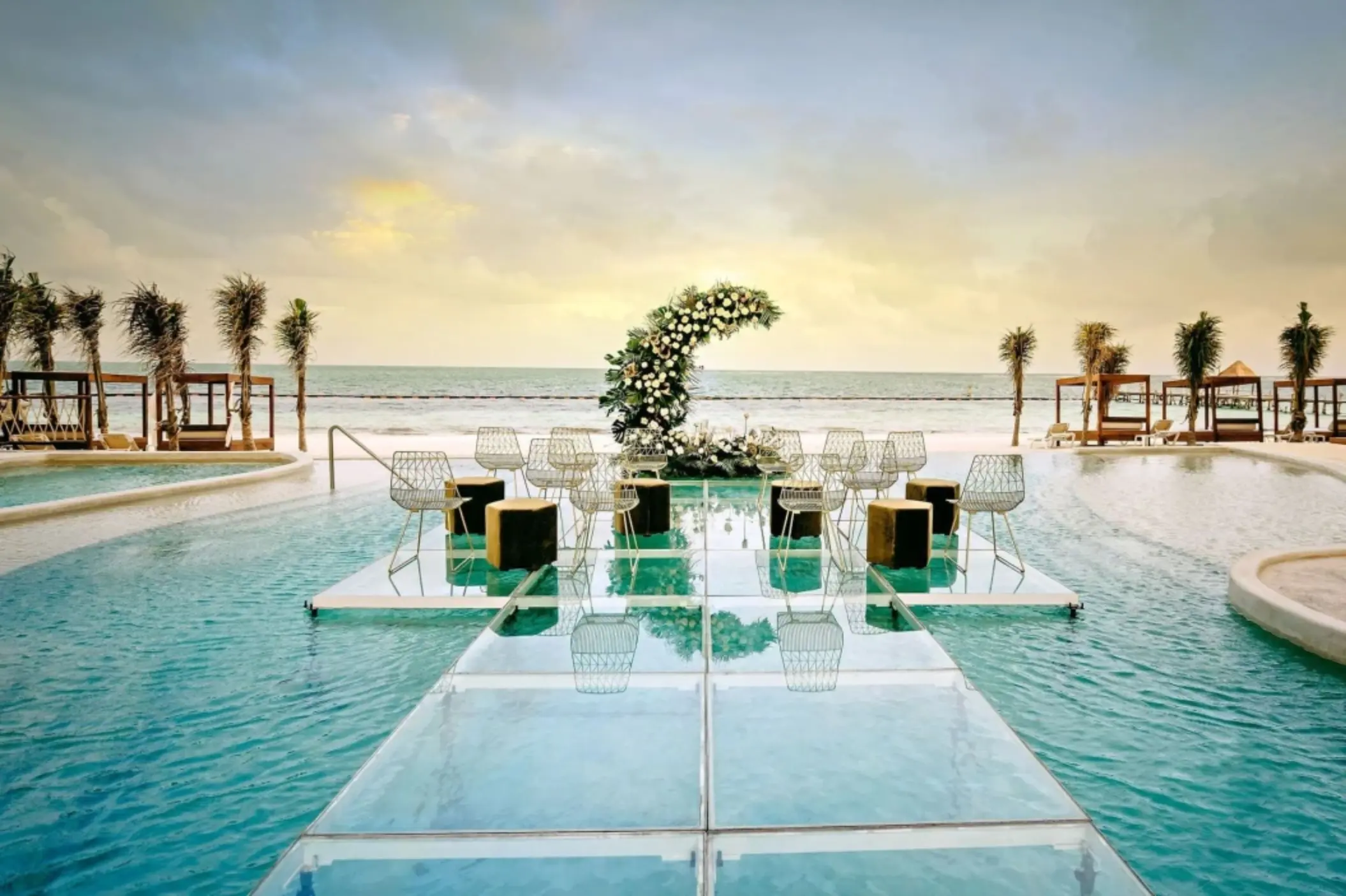 Floating wedding ceremony platform over a pool with ocean backdrop, floral arch, and modern wire chairs.