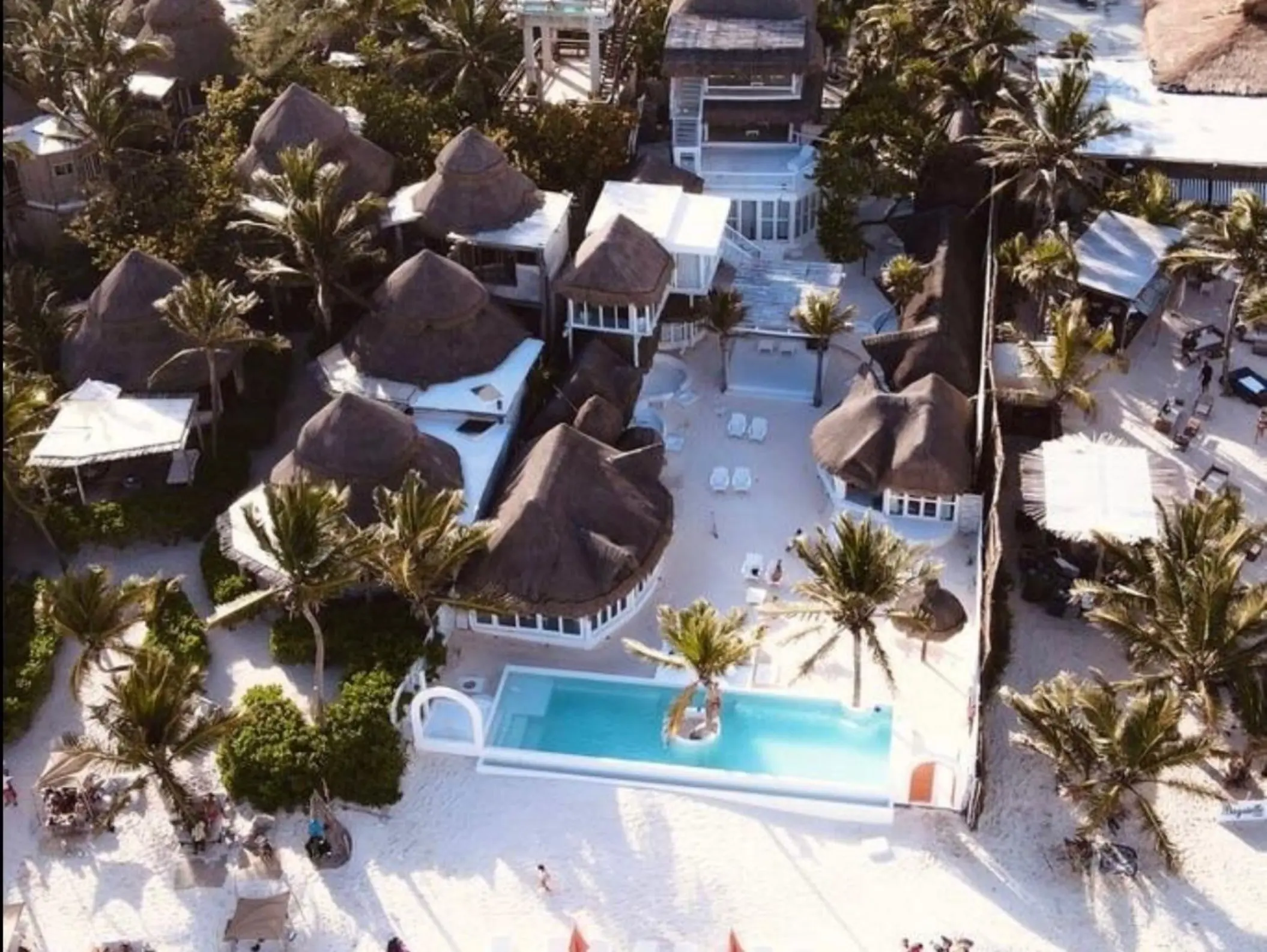 Aerial view of the beach house complex, showing thatched-roof bungalows, a bright pool, and the sandy beach area.