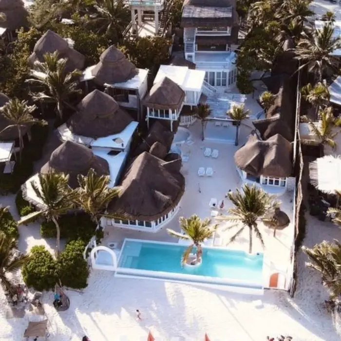 Aerial view of the beach house complex, showing thatched-roof bungalows, a bright pool, and the sandy beach area.