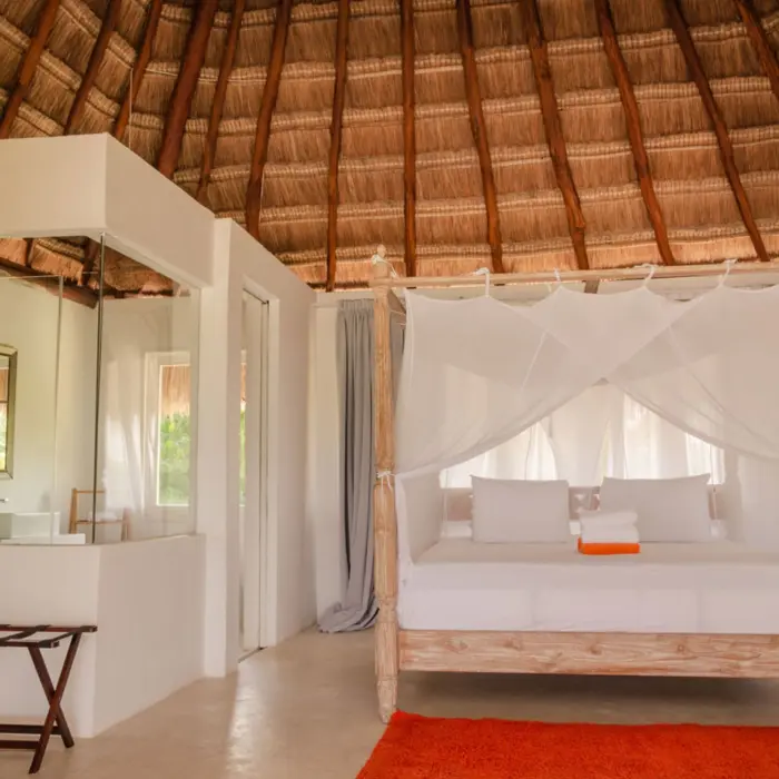 Interior of a casita with a king-sized bed draped in white netting, a thatched roof, and orange rug.