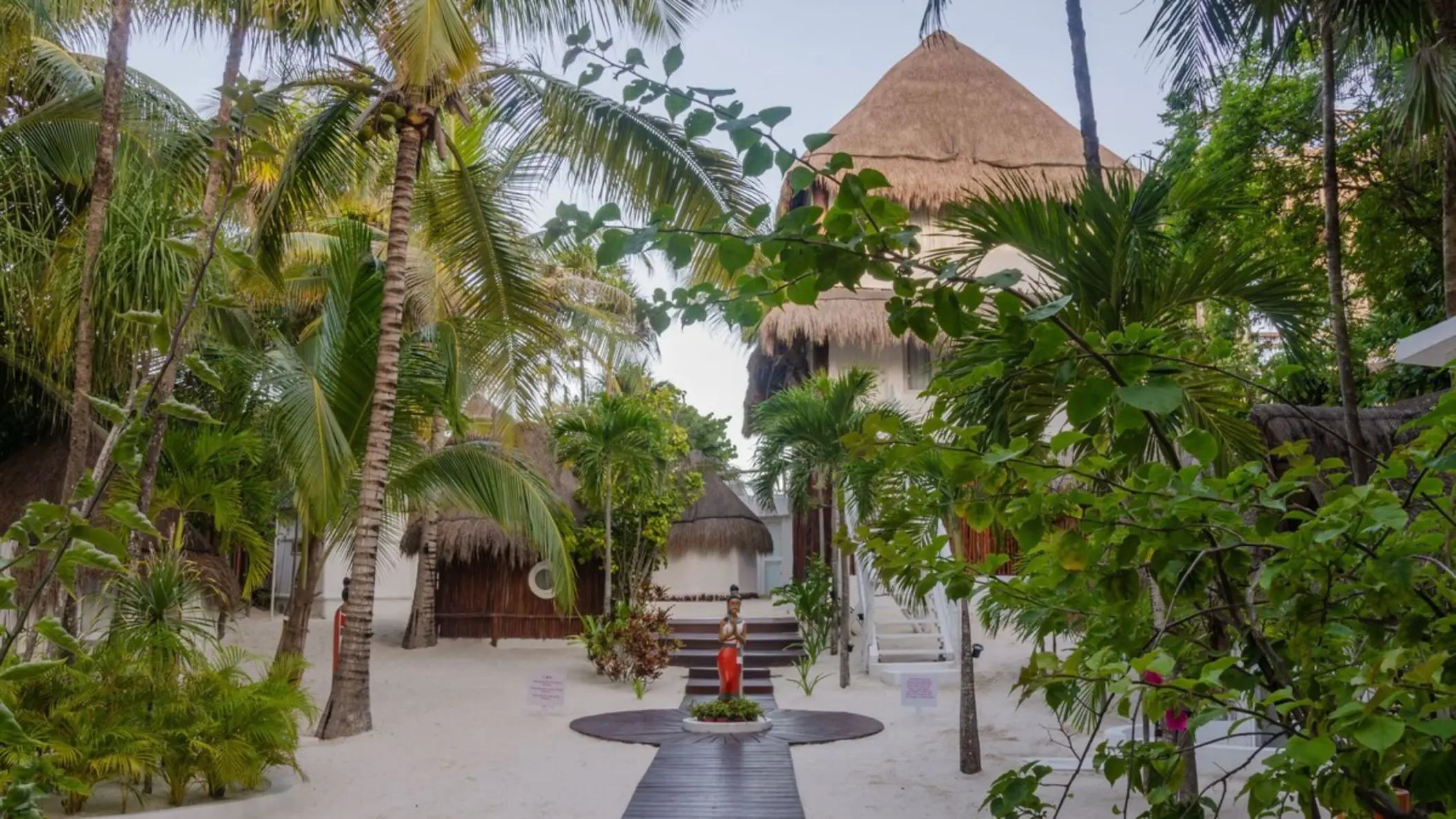 The entrance path through lush greenery and palm trees, leading toward thatched-roof buildings.
